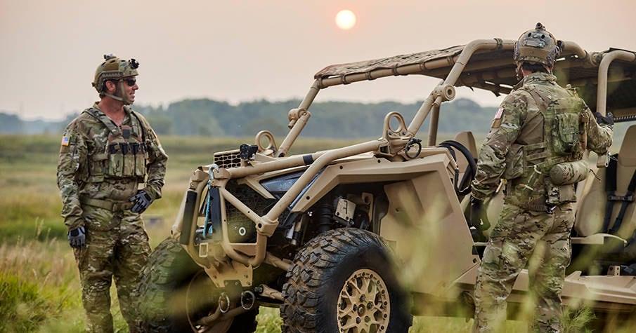 Soldiers standing beside a tan MRZR Alpha in a grassy field at sunset