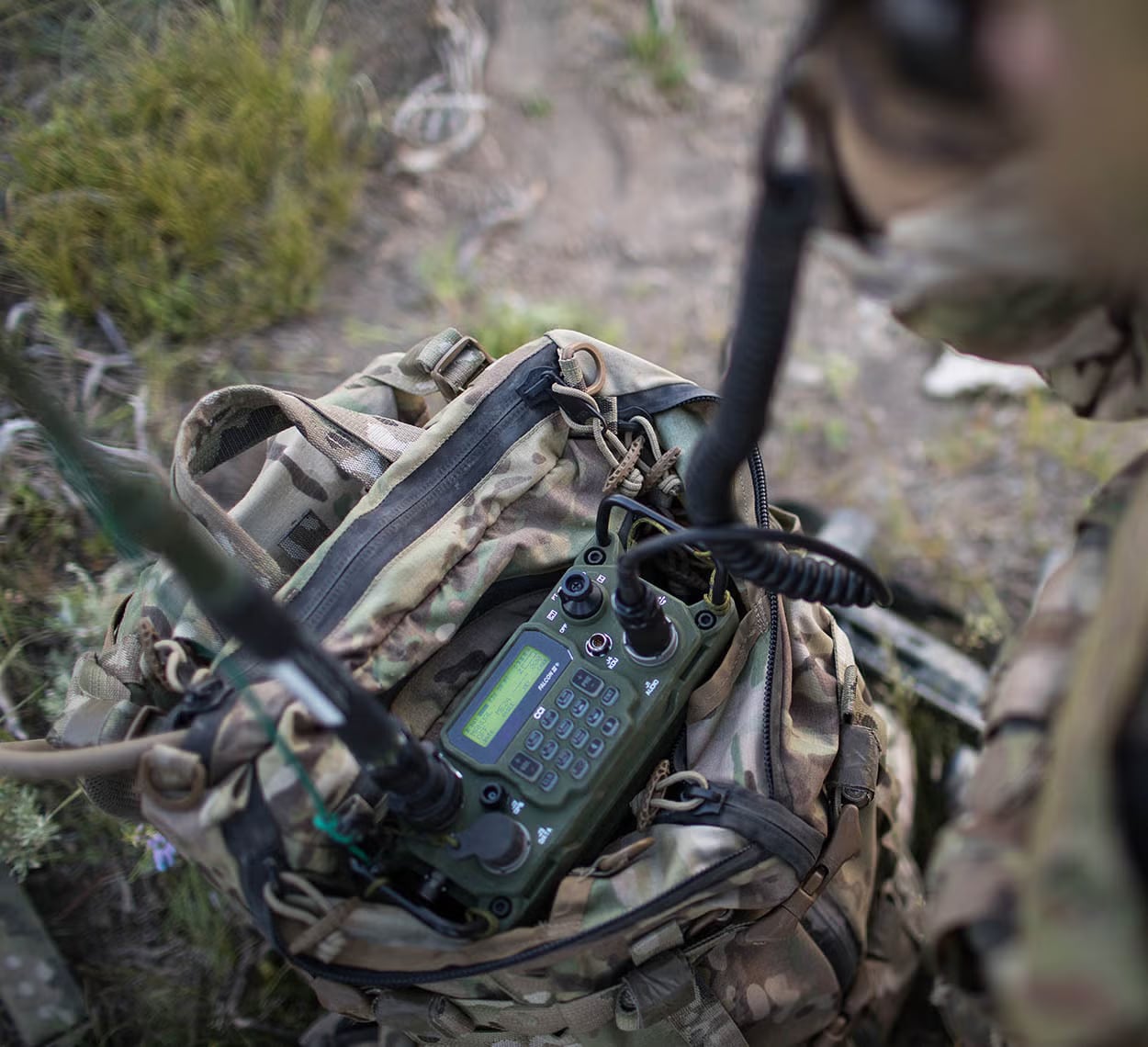 A green HF manpack radio with handset cable attached, sitting on a camouflage pack in dry grass