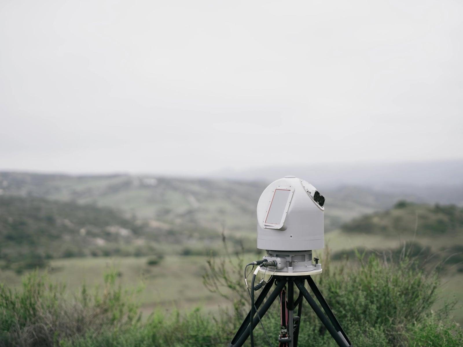 A white dome-shaped sensor on a black tripod on a grassy hillside under an overcast sky